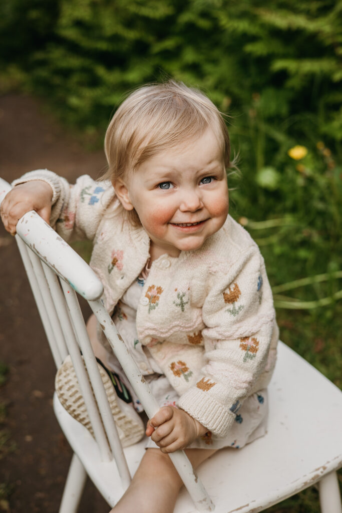 Hymyilevä taapero istuu valkoisella tuolilla puutarhassa – rento miljööperhekuvaus luonnossa