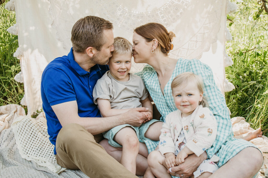 tyttö katsoo kameraan ja vanhemmat ihastelevat isoveljeä perhekuvauksessa