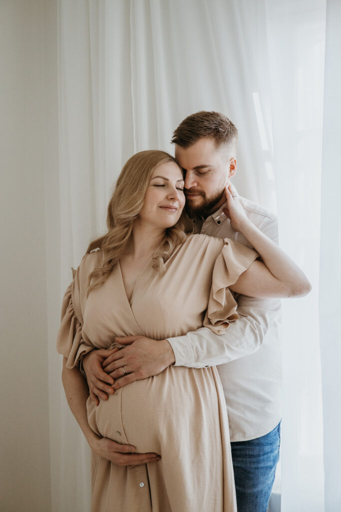 Pariskunta halailee odotusvatsan kanssa ikkunan valossa – studio odotuskuvaus