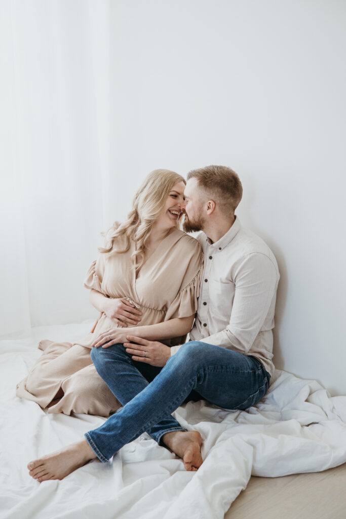 Odottava äiti ja puoliso sylikkäin vaaleassa studiossa – raskauskuvaus luonnonvalossa