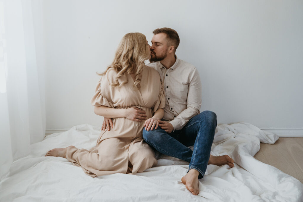 Odottava äiti ja puoliso sylikkäin vaaleassa studiossa – raskauskuvaus luonnonvalossa